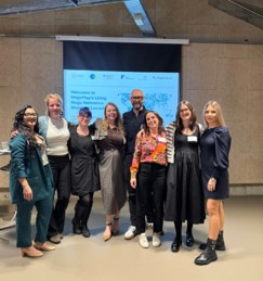 Photograph of eight people standing in front of a white presentation screen displaying a welcome message for a Living Wage event. Individuals are dressed in business casual attire, smiling, and posing in a modern indoor setting with concrete walls and ceiling beams.